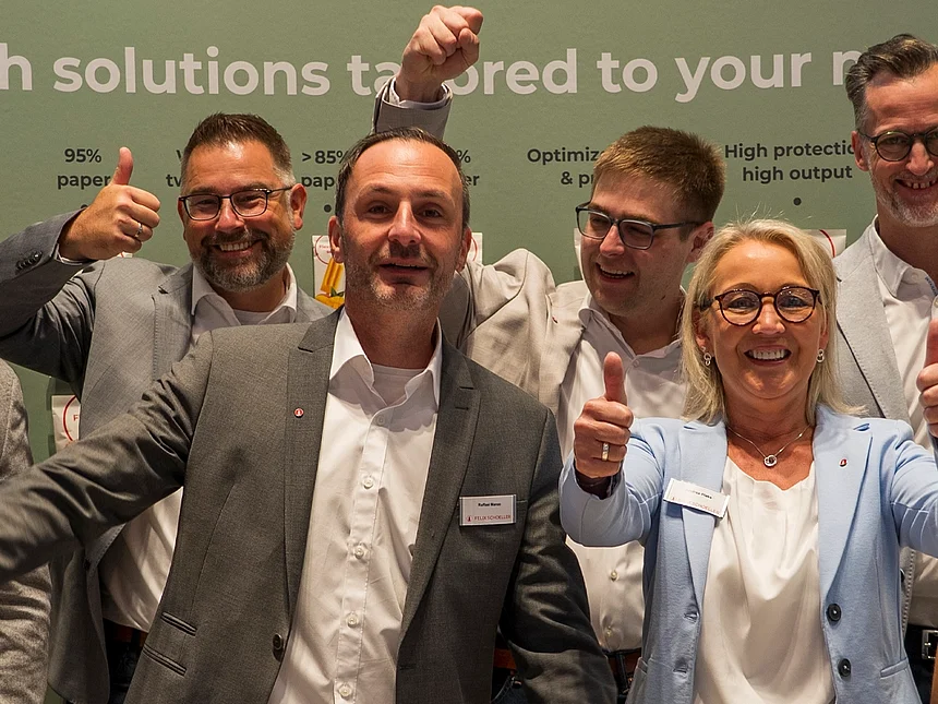 Team von Felix Schoeller jubelt und zeigt Daumen hoch am Messestand vor einer Wand mit dem Slogan „With solutions tailored to your needs“