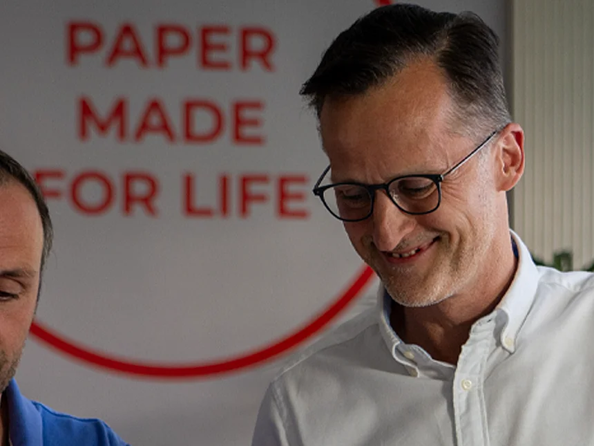 Zwei Männer blicken lächelnd auf ein Dokument vor einer Wand mit dem Markenversprechen „Paper made for life“