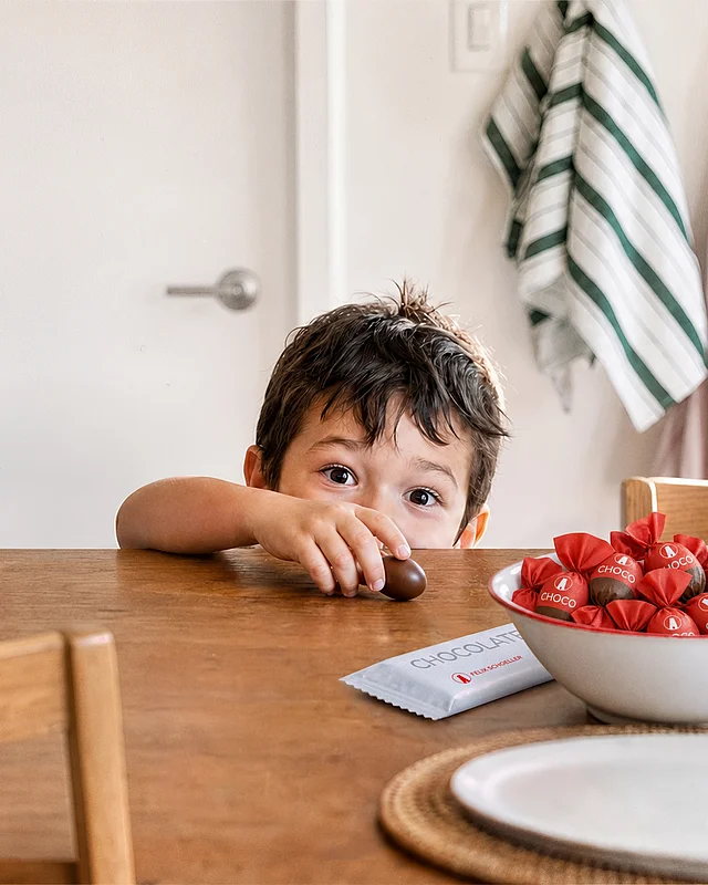 Junge greift am Esstisch nach einer Schokopraline, daneben Schale mit rot papierverpackten Pralinen und Schokoladentafel